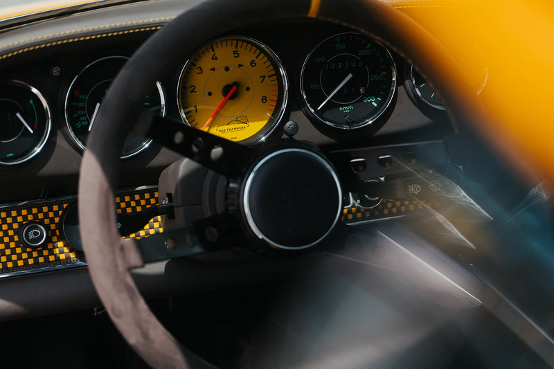 Close-up of a Yellow Triebwerk F4.3 Porsche 911 steering wheel and dashboard with visible gauges.