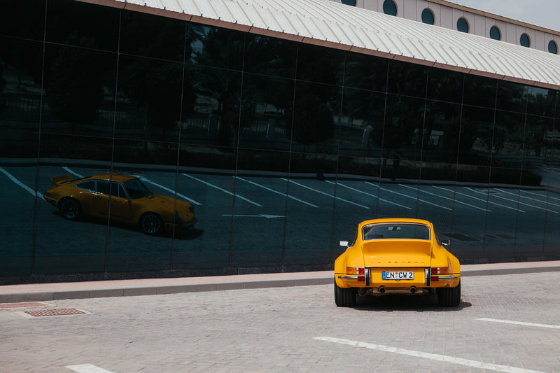 Yellow Triebwerk F4.3 Porsche 911 parked in front of a modern building with reflective glass facade