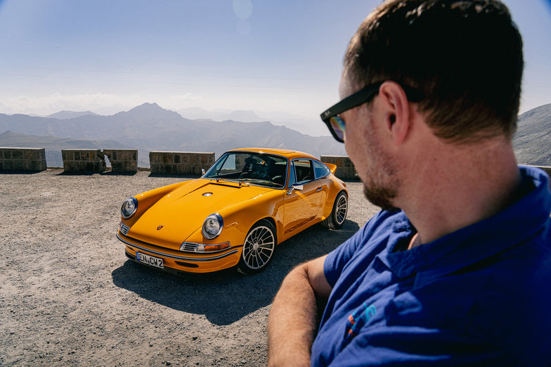 Man in sunglasses looking at a Yellow Triebwerk F4.3 Porsche 911 with mountains in the background